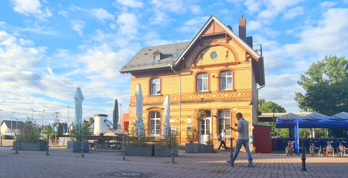 Vor dem alten Bahnhofsgebäude stehen Tische und Stühle. Es laufen Leute von der Treppe zum Parkplatz.